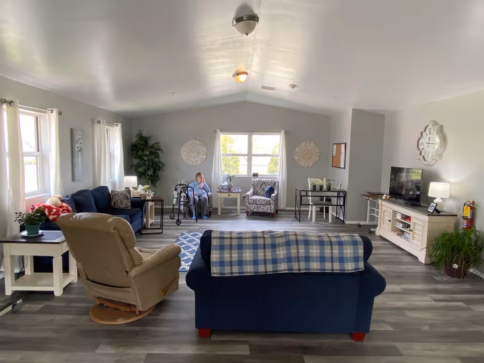 A spacious and well-lit living room in an assisted living facility with multiple seating options including a beige recliner, a blue couch with a plaid blanket, and an armchair. A woman is sitting near the window with a walker beside her. The room has light gray walls, wood flooring, a TV on a white stand, decorative wall art, plants, and several windows with white curtains.