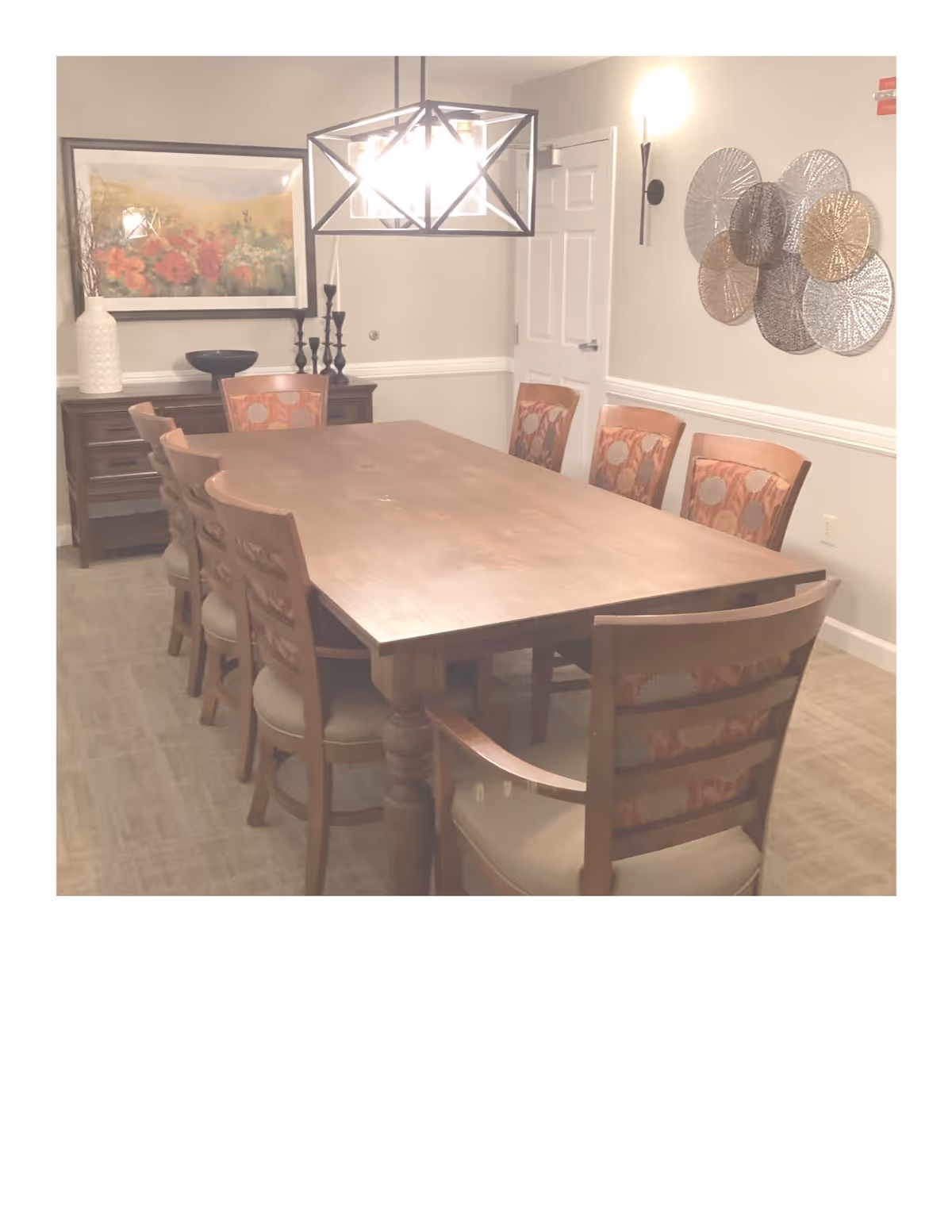 A dining room with a long wooden table surrounded by eight chairs with patterned upholstery. On the left wall, there is a sideboard with decorative items including a large white vase, a black bowl, and black candlesticks. Above the sideboard hangs a framed painting of flowers. A modern geometric chandelier hangs above the table, and a decorative metal wall art piece is mounted on the right wall. The room has beige walls and carpeted flooring.