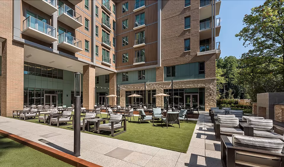 Outdoor seating area at The Seneca Rockville featuring multiple cushioned chairs and tables arranged on artificial grass and paved surfaces, surrounded by a multi-story building with balconies and large windows, with trees visible in the background under a clear sky.