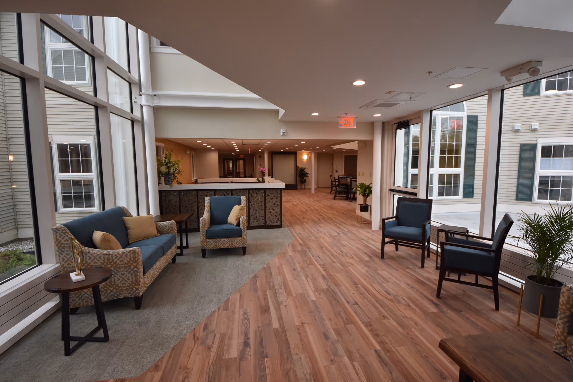 A spacious and well-lit senior living community common area with large windows allowing natural light to fill the room. The space features comfortable seating including a patterned sofa with cushions, armchairs, small side tables, and potted plants. The flooring is a combination of wood and carpet, and the area extends into a hallway with additional seating and decor.