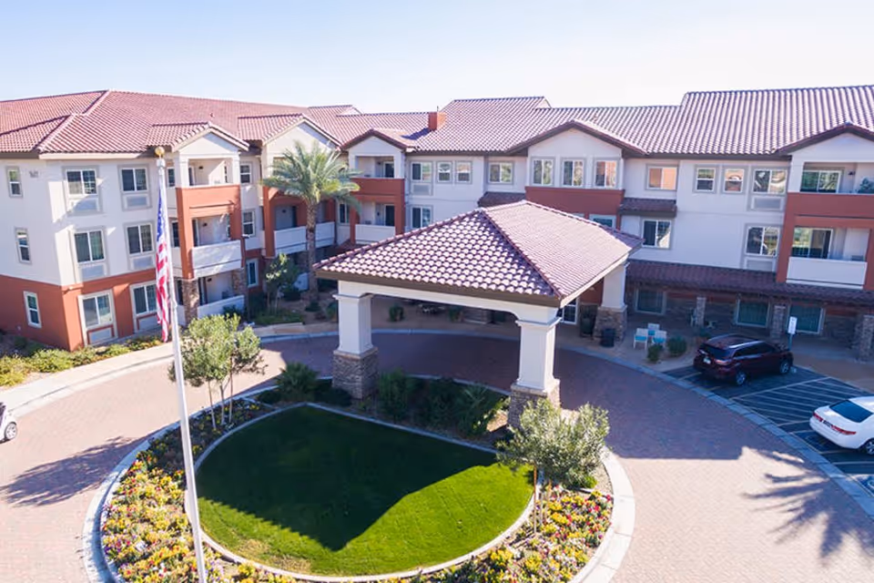 Exterior view of a senior living facility with a circular driveway, landscaped garden with flowers and grass, a flagpole with an American flag, and a covered entrance. The building has three stories with multiple windows and a red-tiled roof.