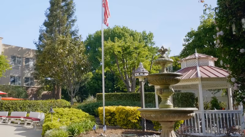 Outdoor courtyard with a multi-tier fountain, gazebo, American flag on a pole, lounge chairs and landscaped gardens in front of the facility building.