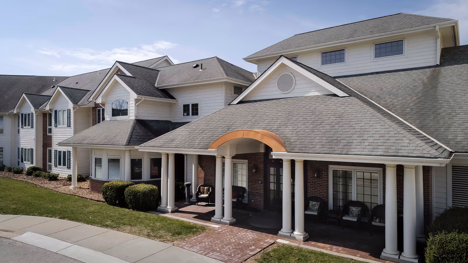Exterior view of a senior living facility building with white siding and brick accents, featuring a covered entrance supported by white columns and outdoor seating. The building has multiple windows and a well-maintained lawn with shrubs.
