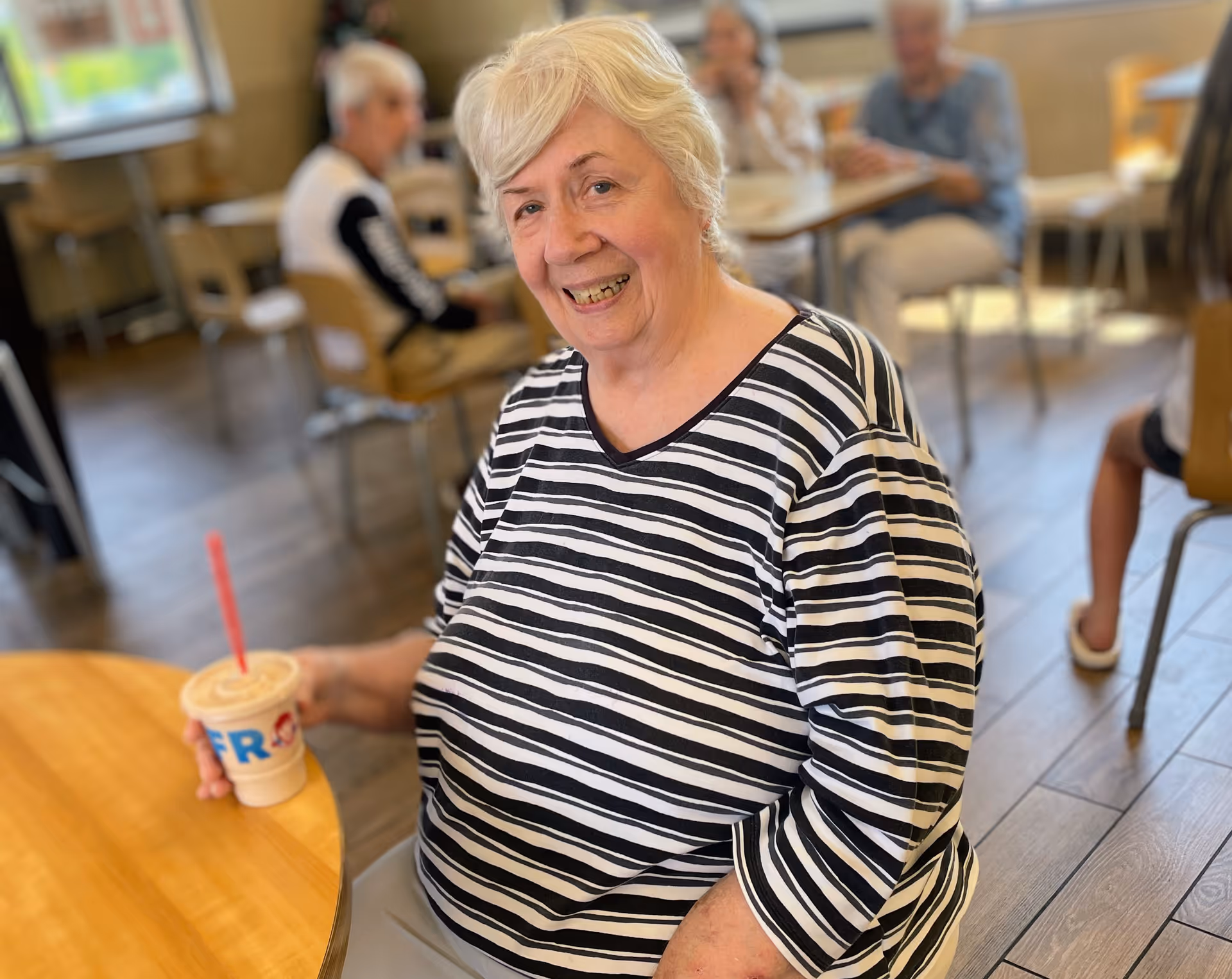 An elderly woman with short white hair wearing a black and white striped shirt is sitting at a wooden table in a dining area, smiling and holding a drink with a red straw. In the background, other elderly people are seated at tables in a well-lit room with wooden flooring.