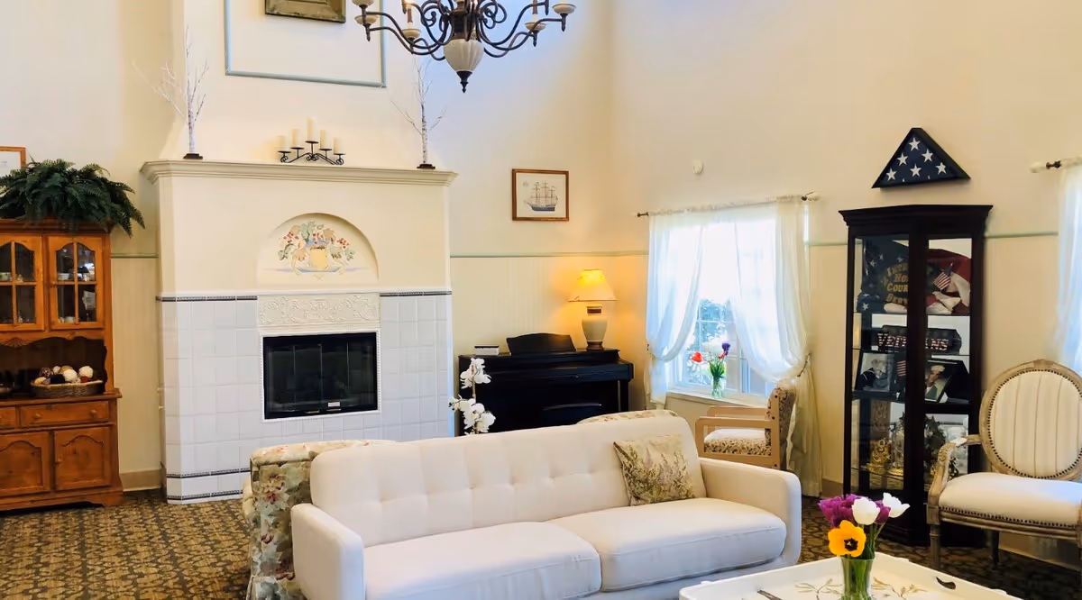 Bright, cozy senior living room with a white sofa facing a tiled fireplace, piano, display cabinet, and seating near curtained windows.