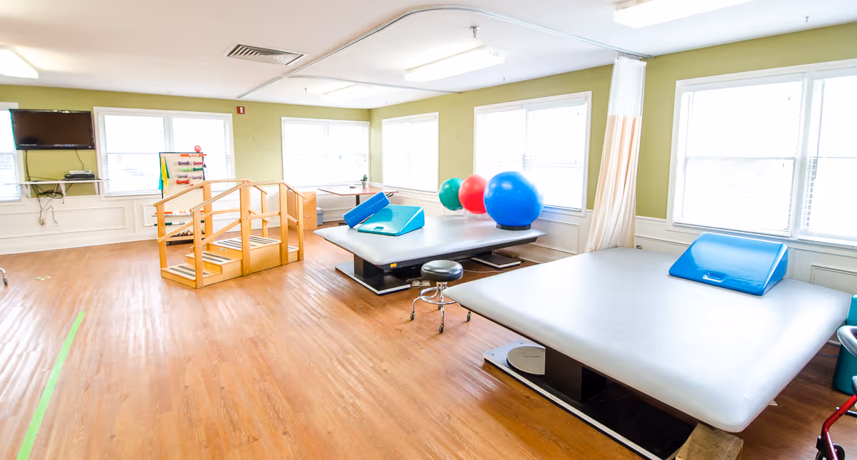 A bright physical therapy room with wooden floors, large windows, and light green walls. The room contains two large padded therapy tables, colorful exercise balls, a small wooden staircase for rehabilitation exercises, a stool, and a television mounted on the wall.