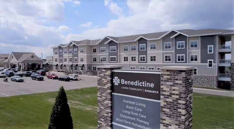 Exterior view of Benedictine Living Community-Bismarck, showing a large multi-story building with numerous windows and a parking lot with several cars. In the foreground, there is a brick sign displaying the facility's name and services including Assisted Living, Basic Care, Long-Term Care, Outpatient Therapy, and Short-Term Rehabilitation.