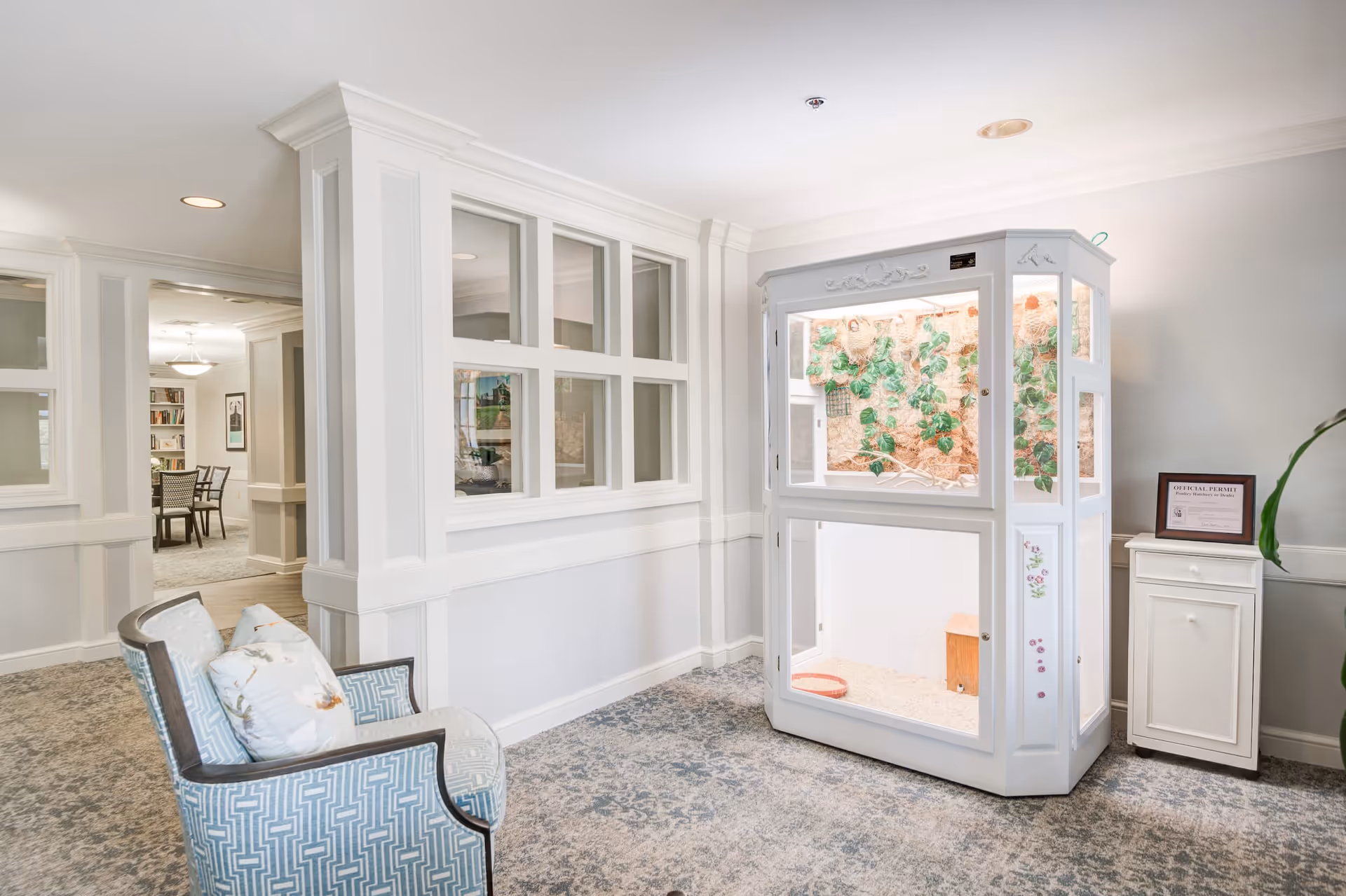 A bright and clean interior space in a senior living facility featuring a large white enclosure with plants inside, a patterned armchair with cushions, and a small white cabinet with a framed certificate on top. The room has light-colored walls, carpeted flooring, and windows looking into an adjacent room with a dining area visible.