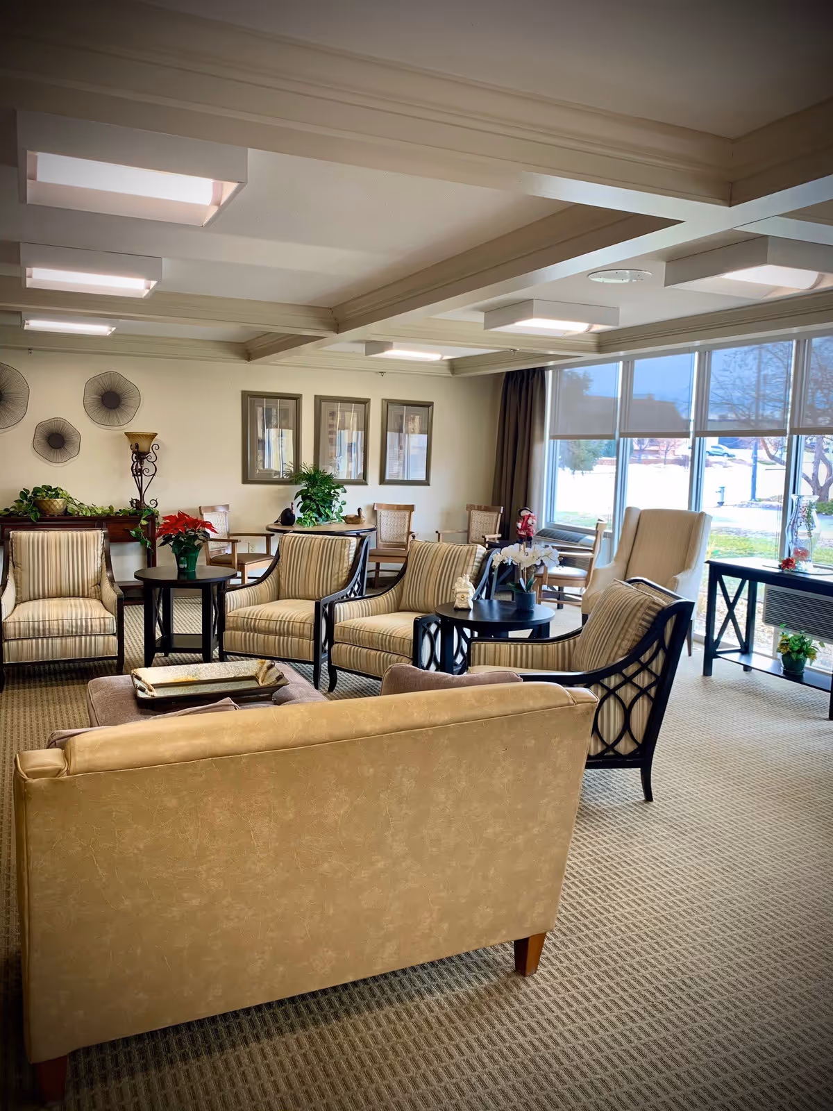 A well-lit living room area in an assisted living center featuring multiple cushioned armchairs and a sofa arranged around small round tables. The room has large windows with a view of the outdoors, framed pictures on the wall, decorative plants, and a carpeted floor.