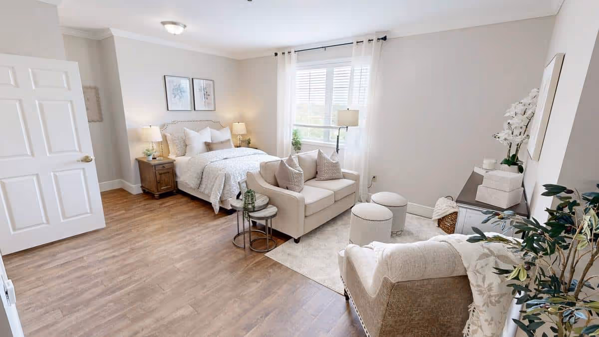 A bright and spacious bedroom with a large bed, two bedside tables with lamps, a small beige loveseat with pillows, two round ottomans, an armchair, a window with white curtains, and decorative plants and artwork on the walls.