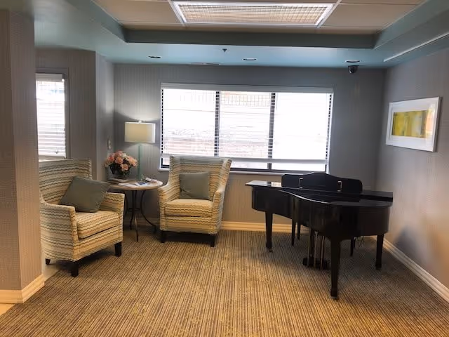A cozy sitting area with two striped armchairs and a small round table with a lamp and a vase of flowers between them. A black grand piano is positioned near a large window with blinds. The room has light-colored walls, carpeted floor, and a framed artwork on the wall.