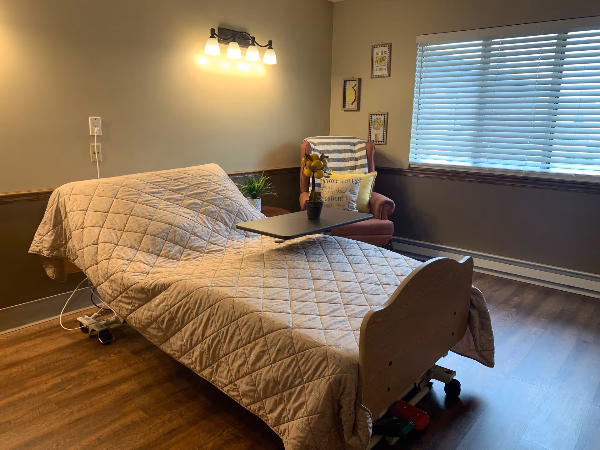 A room with a hospital-style adjustable bed covered with a beige quilted blanket. Next to the bed is a small table with a potted plant on it. In the corner, there is a comfortable armchair with a striped throw and decorative pillows. The room has wooden flooring, a window with blinds, and a wall-mounted light fixture above the bed.