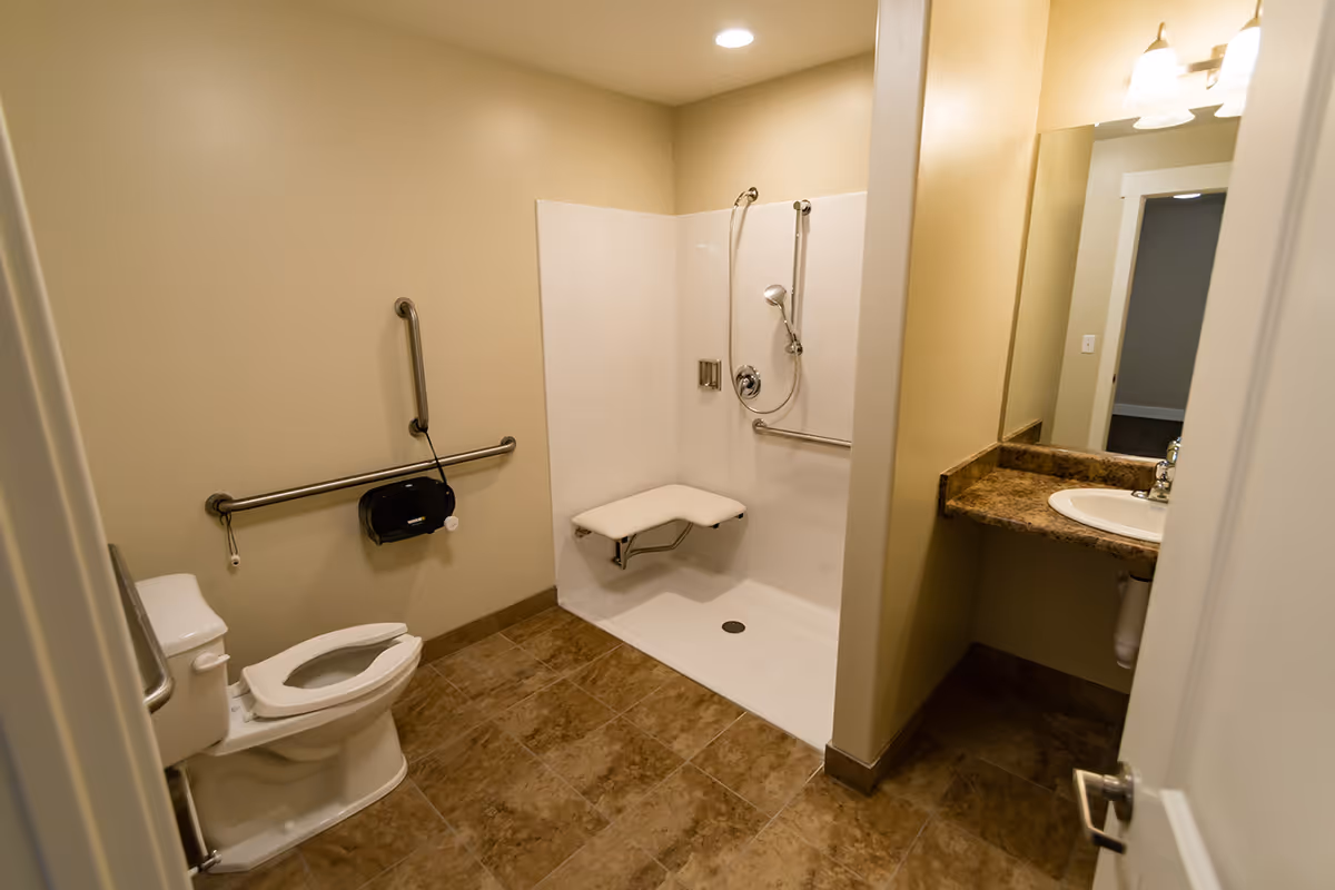 A clean and accessible bathroom with beige walls and brown tiled floor. The bathroom features a white toilet with grab bars on the walls, a walk-in shower with a fold-down seat and handheld showerhead, and a sink with a granite countertop and a mirror above it.