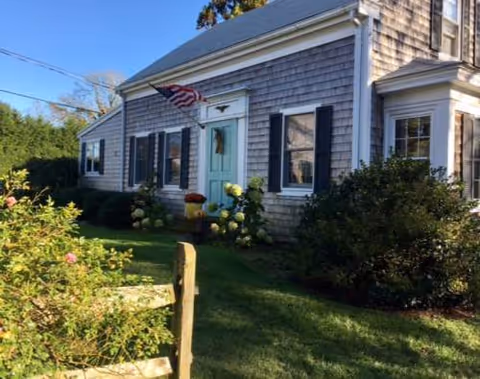 Shingle-clad senior living facility exterior with turquoise door, American flag, and manicured garden.