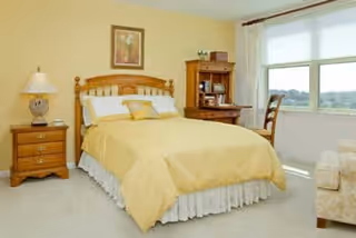 A bright bedroom with a wooden bed frame and yellow bedding, a wooden nightstand with a lamp, a wooden desk with shelves, a chair by a large window with white curtains, and a beige armchair partially visible.