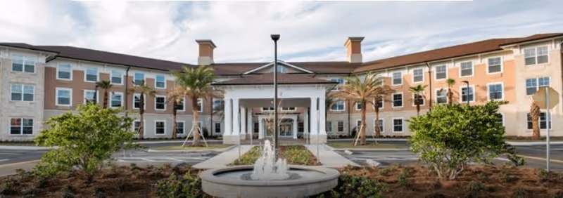 Front exterior view of Canterfield of Tallahassee, a three-story senior living facility with a central entrance featuring white columns, surrounded by palm trees and landscaping with a water fountain in the foreground.