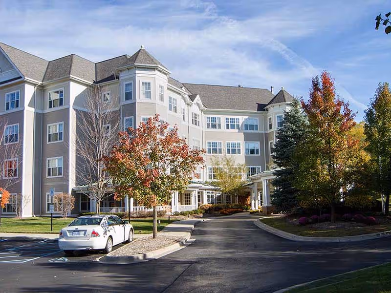 Exterior view of a multi-story senior living facility building with beige and white siding, surrounded by trees with autumn foliage and a parking lot with a white car parked in a handicapped space.