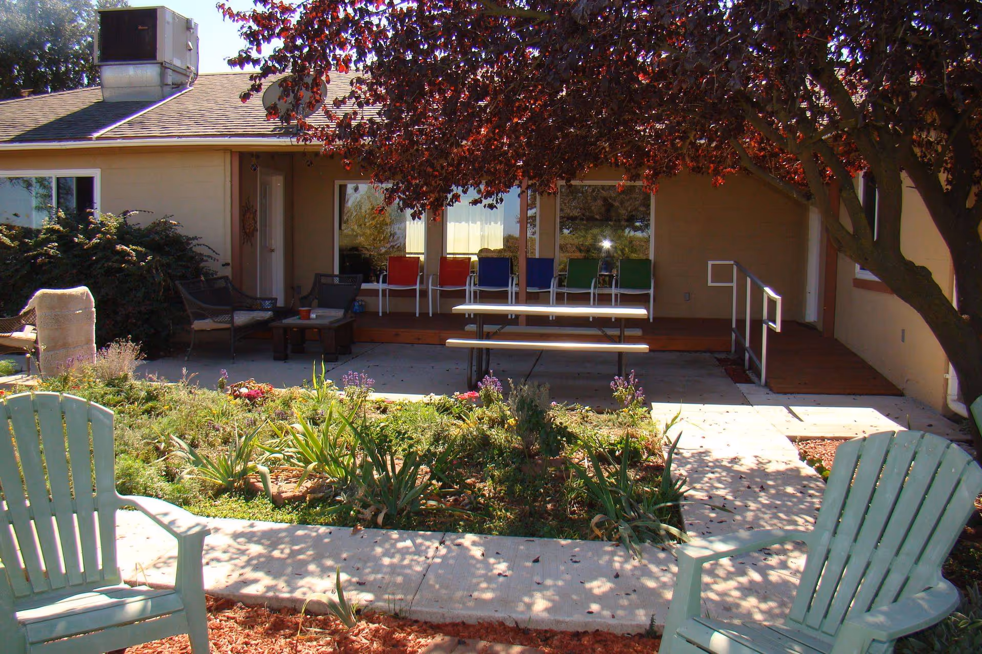 Outdoor patio area at Orchard View Senior Care featuring a garden with various plants and flowers, two light green Adirondack chairs in the foreground, a concrete walkway, and a covered porch with colorful chairs and a picnic table. A tree with red leaves provides shade over part of the patio.