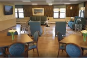 Sunlit common lounge with teal chairs, tables, flowers, a sofa, and a wall-mounted TV.
