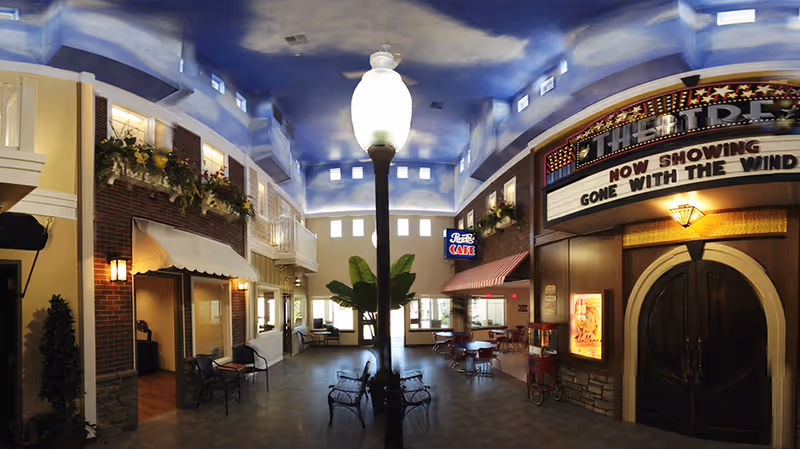 Indoor senior living common area styled like a small-town street with a central lamp post, seating, a cafe sign, and a theater marquee.