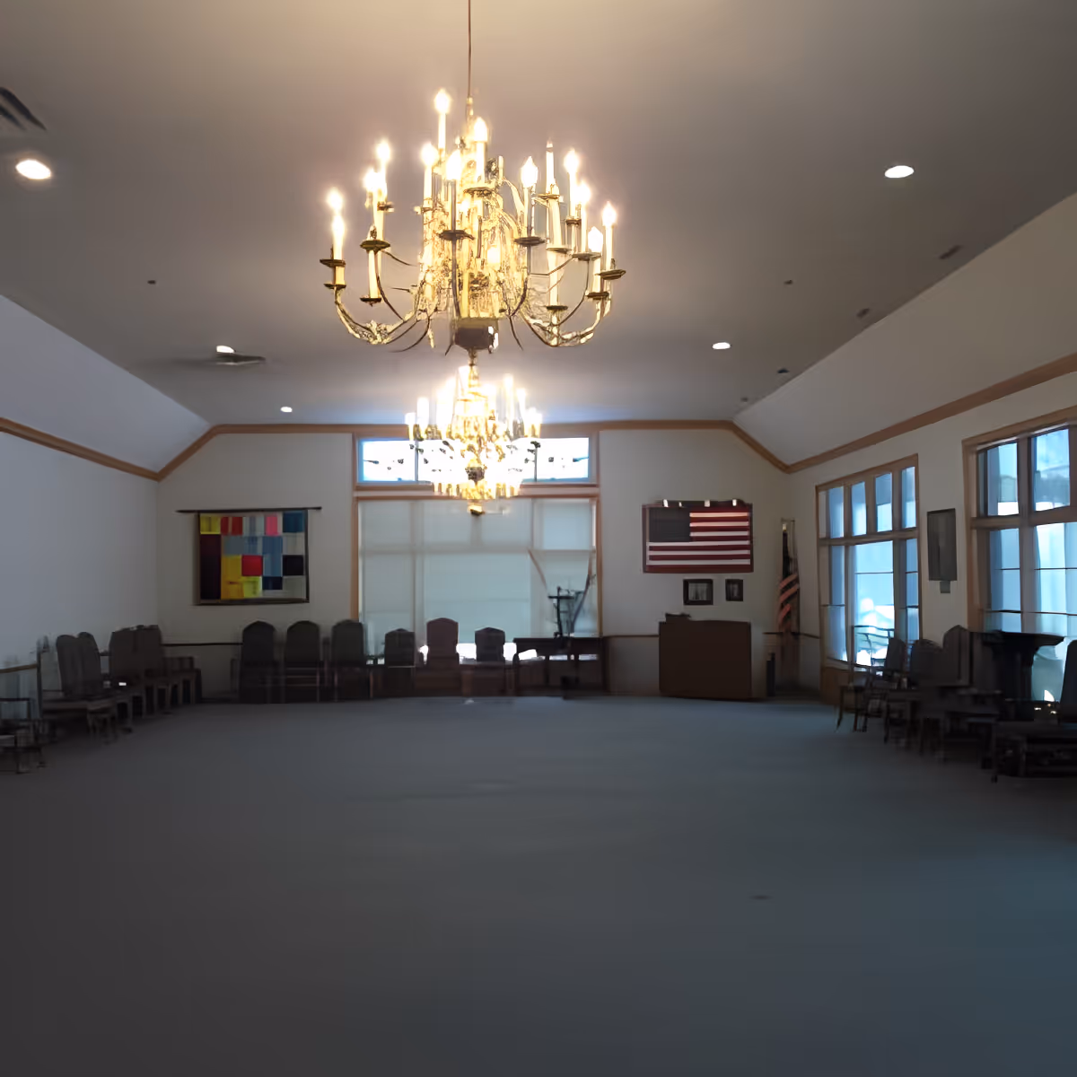 A large, empty room with rows of chairs lined up against the walls on both sides. The room features two ornate chandeliers hanging from the ceiling, large windows on the right side, and an American flag displayed on the far wall. The walls are white with wooden trim, and there is a colorful quilt hanging on the left wall.