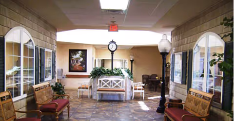 Indoor hallway or common area with tiled floor, wooden benches with red cushions on both sides, large windows with white frames, potted plants, a central planter with greenery, and vintage-style street lamps. A clock is mounted on a post in the center of the planter. The walls are beige with a brick pattern, and there is a framed picture on the far wall.