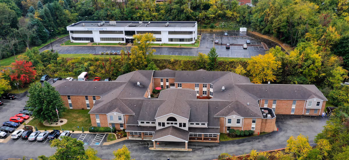 Aerial view of Independence Court of Monroeville, a large brick building with a complex roof structure surrounded by trees with autumn foliage. There are parking lots with several cars parked around the building and a smaller white building behind it. The area is lush with greenery and trees.