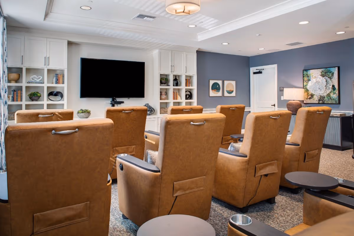 A cozy living room area with multiple brown leather recliner chairs arranged in rows facing a large flat-screen TV mounted on a white wall. The room features built-in white shelves with decorative items and books, a blue accent wall with framed artwork, a door, and a lamp on a side table. The ceiling has recessed lighting and a modern light fixture.