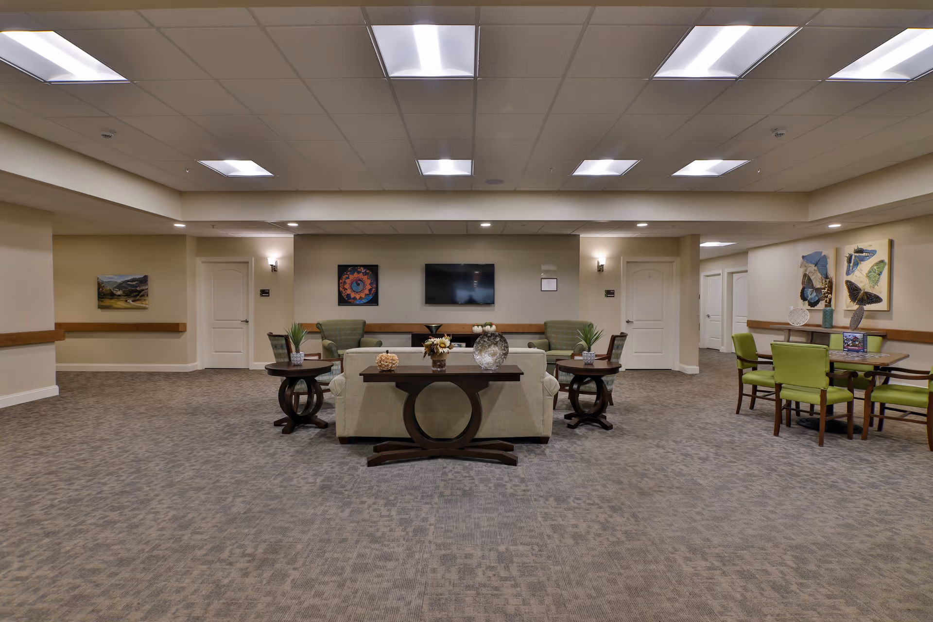 A spacious senior living common area with beige walls and carpeted floor. The room features a central seating arrangement with a beige sofa, two green armchairs, and two round wooden side tables with decorative items. A flat-screen TV is mounted on the far wall, flanked by two doors. To the right, there is a wooden table with four green chairs and butterfly artwork on the wall above it. The ceiling has recessed lighting and several fluorescent light panels.