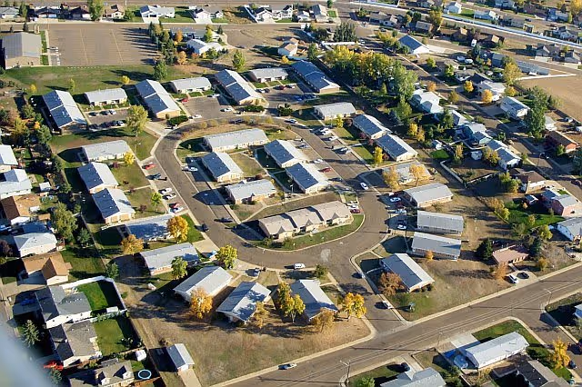 Aerial view of a residential complex with multiple single-story buildings arranged in a circular pattern with roads and parking areas, surrounded by trees and other houses.