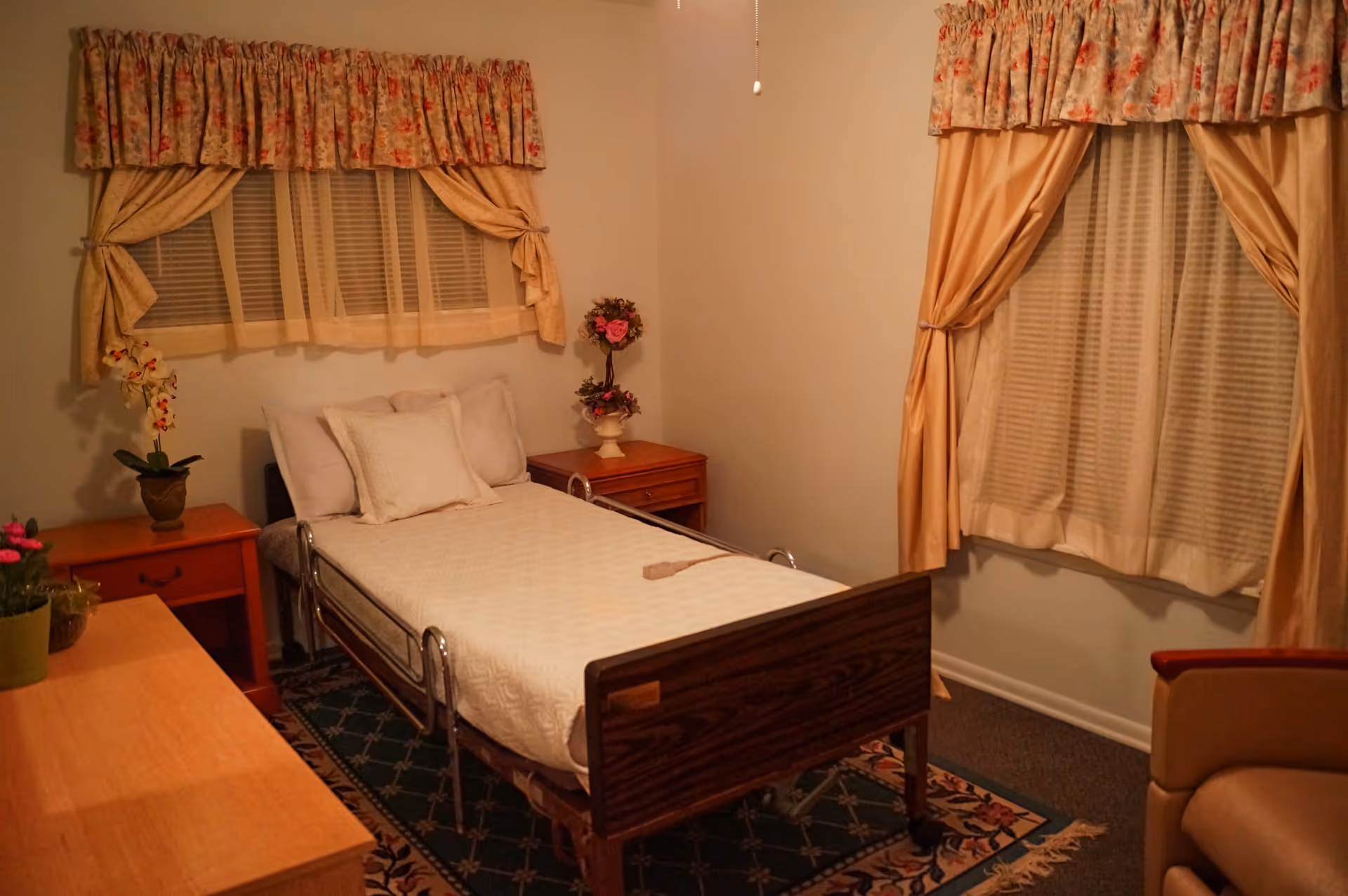 A small bedroom with a single hospital-style bed covered with white bedding and pillows. The room has two windows with floral and beige curtains, a wooden nightstand with a flower vase, a wooden dresser with potted plants, and a beige armchair. The floor has a patterned rug.