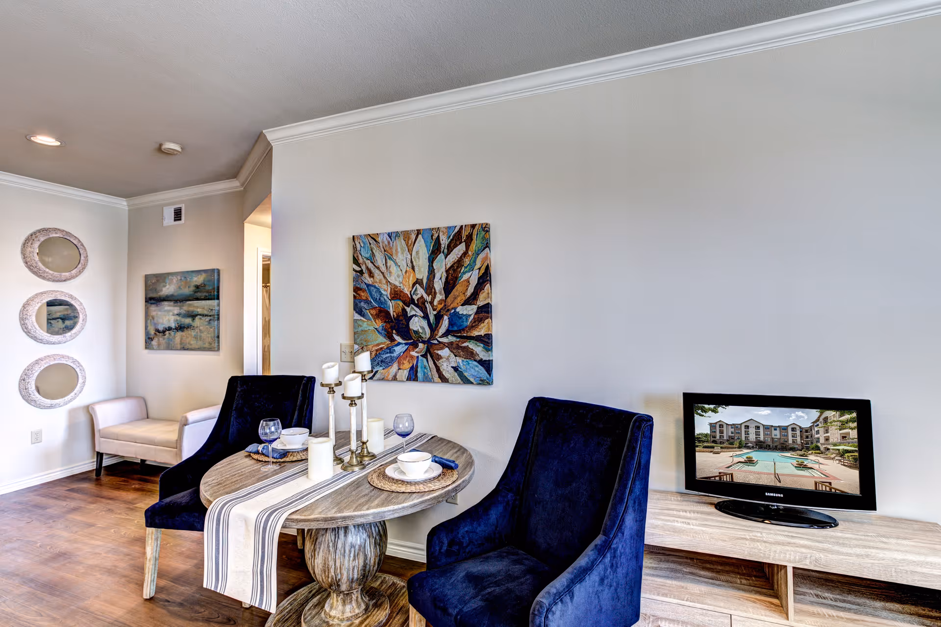 A cozy interior space featuring a small round wooden dining table with a striped table runner, two dark blue upholstered chairs, and table settings including bowls, glasses, and candles. On the wall above the table is a colorful abstract painting. To the left, there is a small beige bench under three decorative round mirrors and a landscape painting. On the right, a wooden console table holds a flat-screen TV displaying an image of an apartment complex with a pool.