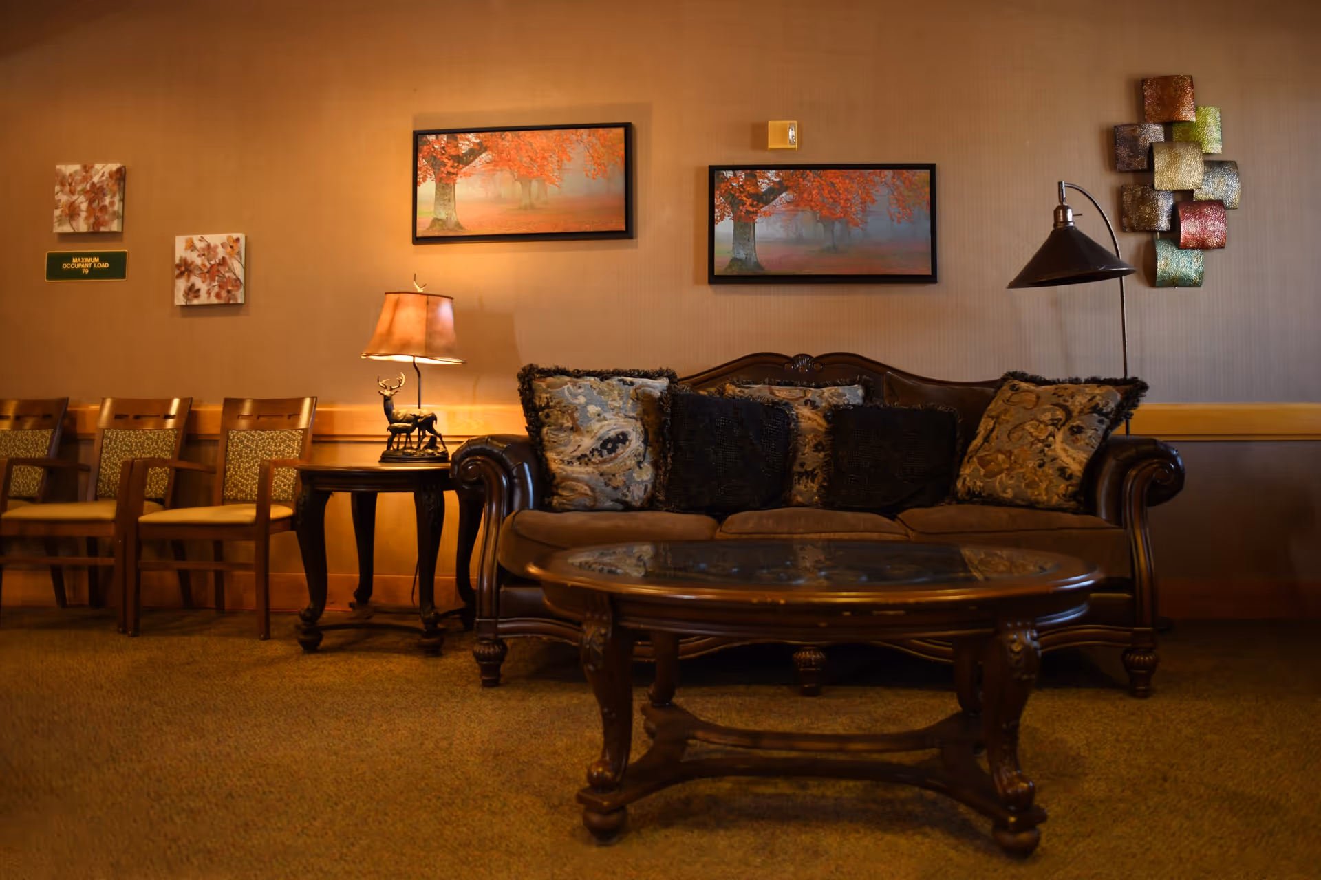 A cozy living room area with a brown leather sofa adorned with patterned cushions, a wooden coffee table with a glass top in front, and two side tables each with a lamp. The wall behind the sofa features two framed pictures of autumn trees with red leaves and decorative wall art. To the left, there are three wooden chairs with patterned cushions lined up against the wall.