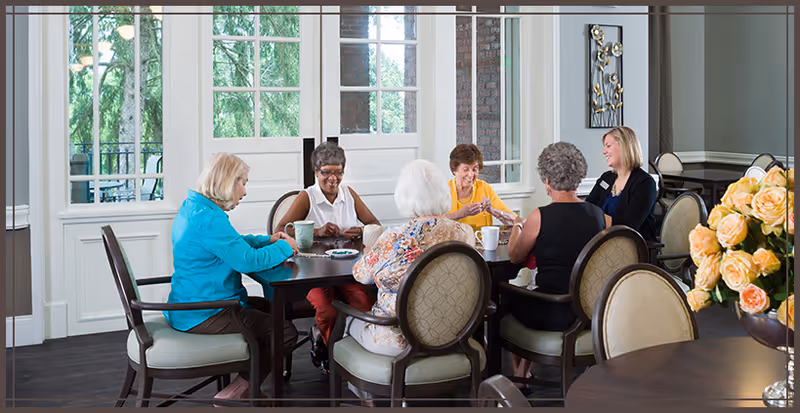 A group of senior women sitting around a dining table in a bright common room, chatting and enjoying drinks.
