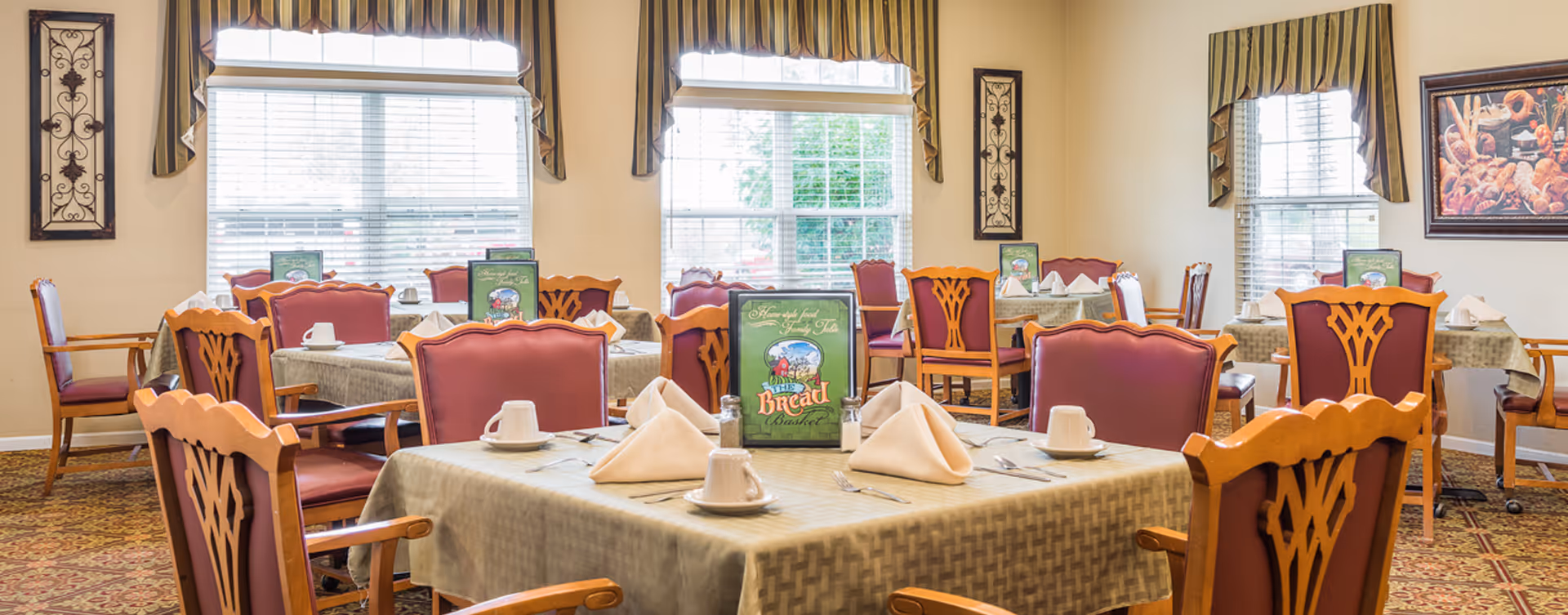 A dining room with multiple tables covered in beige tablecloths, each set with white napkins, cups, and silverware. The chairs have wooden frames with red cushioned seats and backs. Large windows with striped valances allow natural light to fill the room. Decorative wall art and framed pictures adorn the walls.