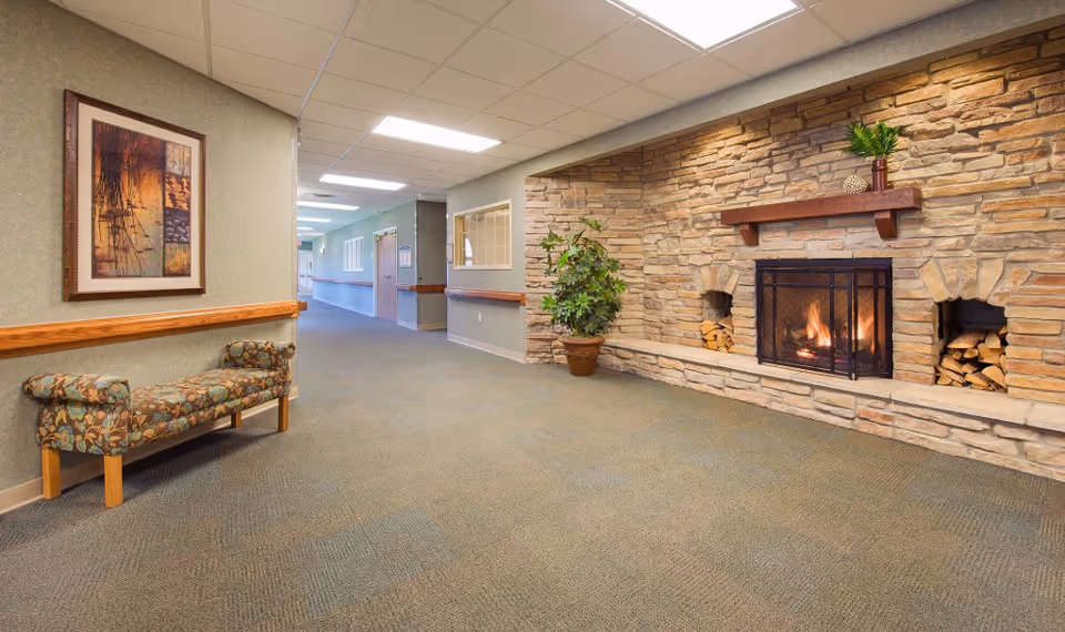 A hallway in a senior living facility with a stone fireplace on the right wall, a potted plant beside it, and a cushioned bench with floral upholstery on the left wall. The hallway is carpeted and well-lit with ceiling lights, and there is a framed abstract painting above the bench.