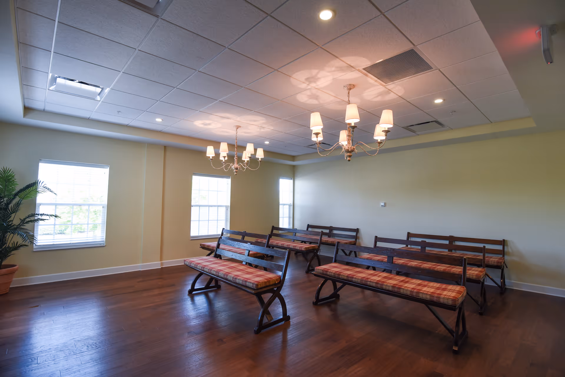 A room with wooden floors and pale yellow walls featuring several wooden benches with plaid cushions arranged in rows. The room is illuminated by two chandeliers with multiple lampshades hanging from the ceiling. There are three windows on one wall allowing natural light to enter, and a potted plant is visible in the corner.