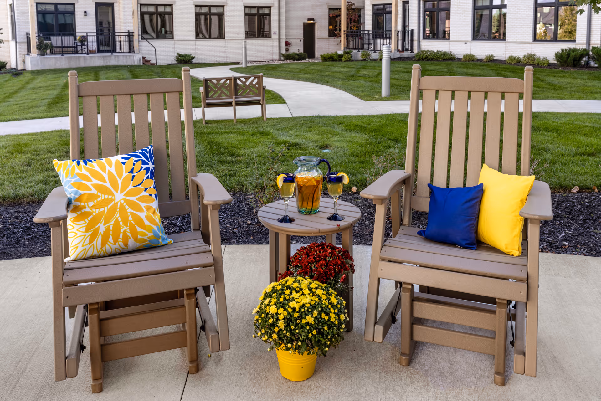Two beige wooden rocking chairs with colorful cushions, one with a yellow and blue floral pillow and the other with solid blue and yellow pillows, are placed on a concrete patio. Between the chairs is a small round table holding a pitcher of iced tea and two glasses with lemon slices. In front of the table are two pots of flowers, one with yellow blooms and the other with red blooms. The background shows a well-maintained lawn, a curved concrete pathway, and a white brick building with large windows.