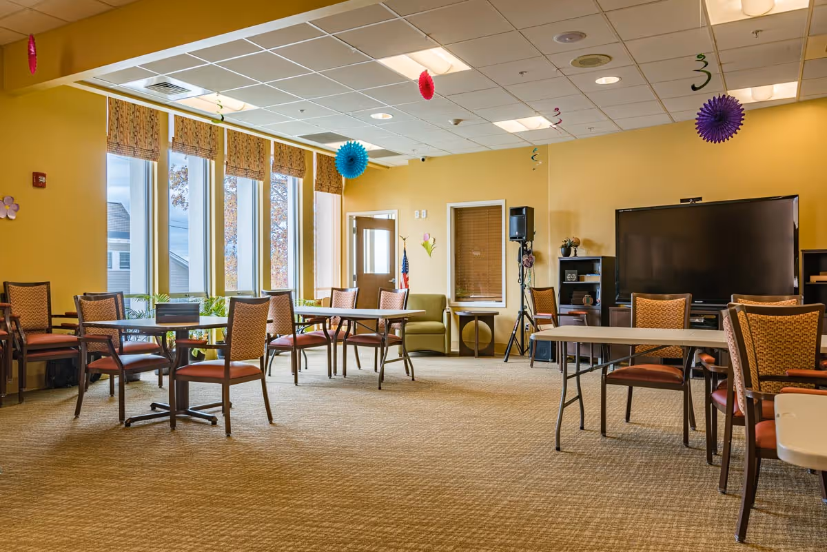 A spacious room with several tables and chairs arranged around the perimeter. The walls are painted yellow, and there are large windows with patterned valances letting in natural light. The ceiling has recessed lighting and hanging colorful decorations. A large flat-screen TV is mounted on a stand against one wall, with shelves and speakers nearby. There is also a green armchair and a door with a window in the background.