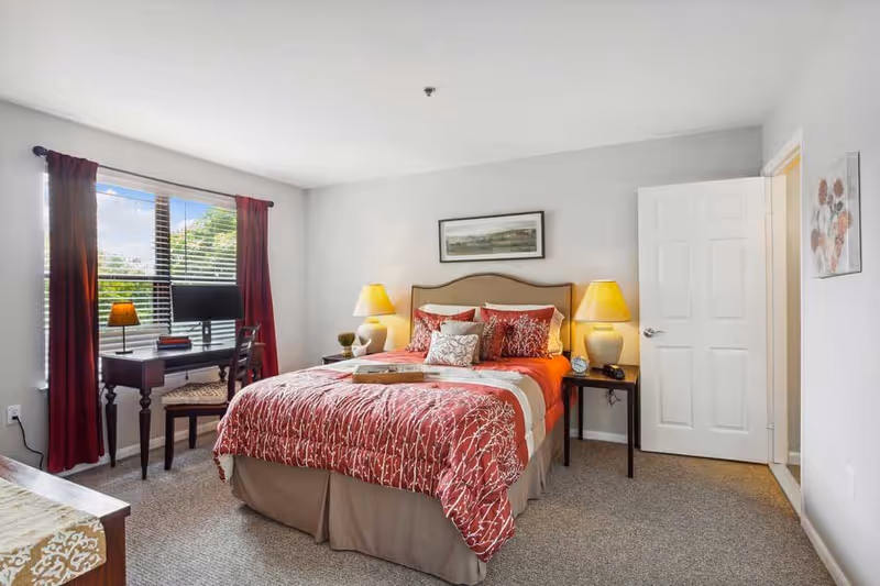 A well-lit bedroom with a bed dressed in red and beige bedding, two bedside tables each with a lamp, a desk with a chair and a lamp near a window with red curtains, and a door open to another room. The walls are light gray and there is a framed picture above the bed.