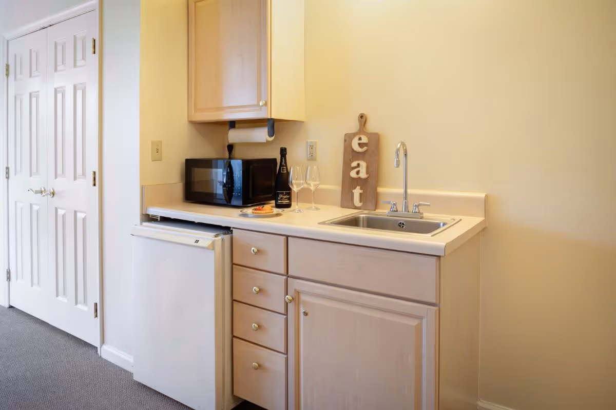 Small kitchenette area with light wood cabinets, a white mini refrigerator, a microwave, a sink with a faucet, and a wooden cutting board with the word 'eat' on it. There are two empty wine glasses and a bottle on the counter along with a small plate of food. A white door is visible to the left.