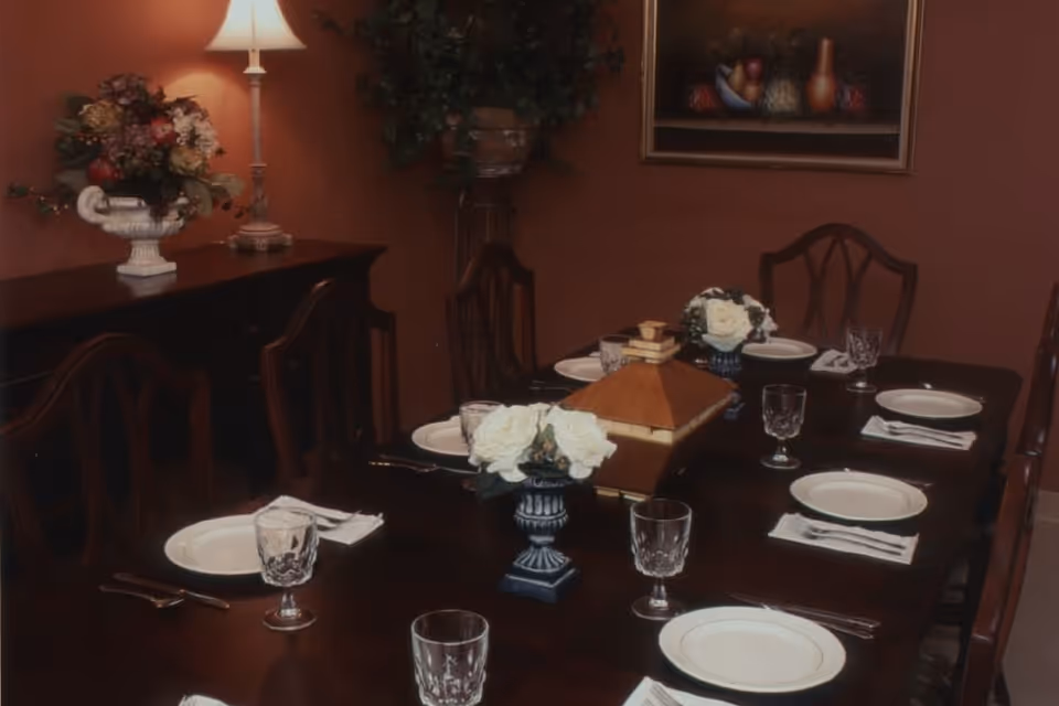 A formal dining room with a dark wooden table set for six people. Each place setting includes a white plate, a folded napkin with silverware, and a clear glass. The table has two small floral centerpieces with white flowers and a decorative wooden box in the middle. In the background, there is a sideboard with a lamp and a floral arrangement, a potted plant on a stand, and a framed painting on the wall.