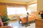 A bright and clean patient room in a healthcare facility featuring a hospital bed with an orange bedspread, an overbed table, a window with patterned curtains, a wooden dresser with a flower arrangement, a green plant, and a comfortable chair.