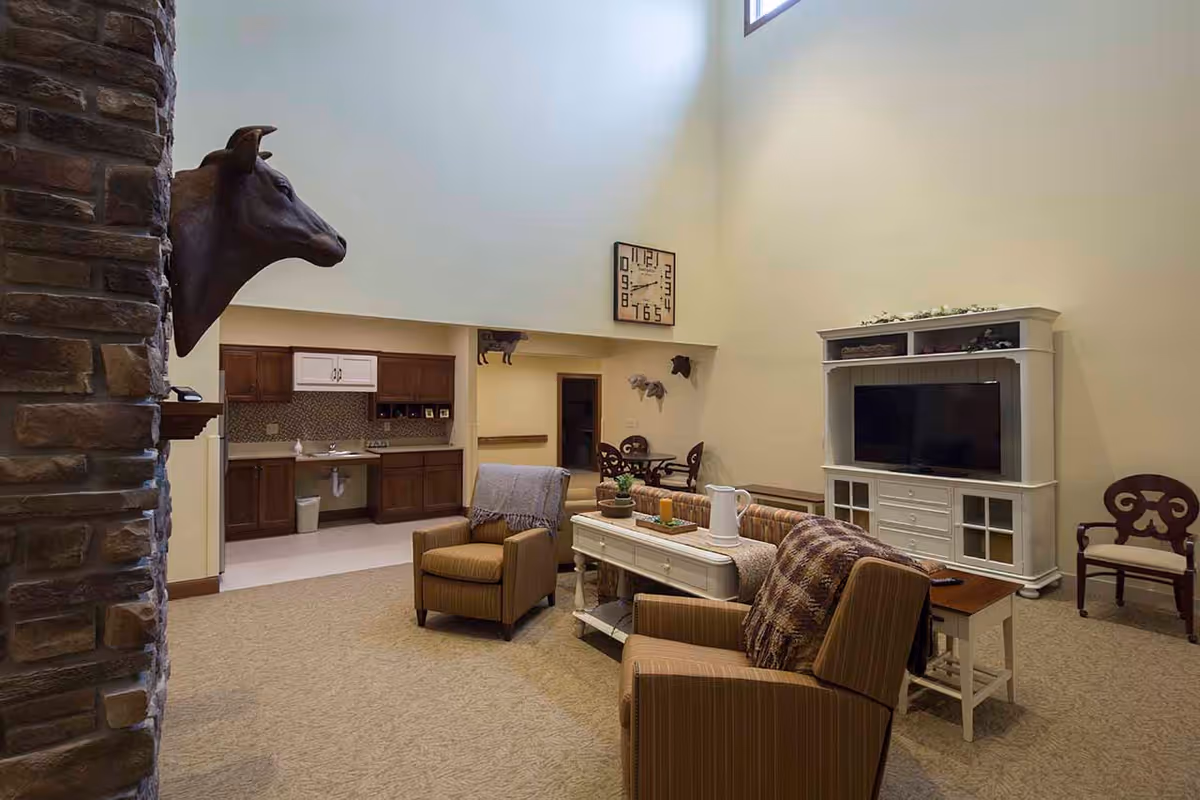 A spacious senior living common area with armchairs around a coffee table, a TV cabinet, a kitchenette in the back, and a mounted animal head on a stone pillar.