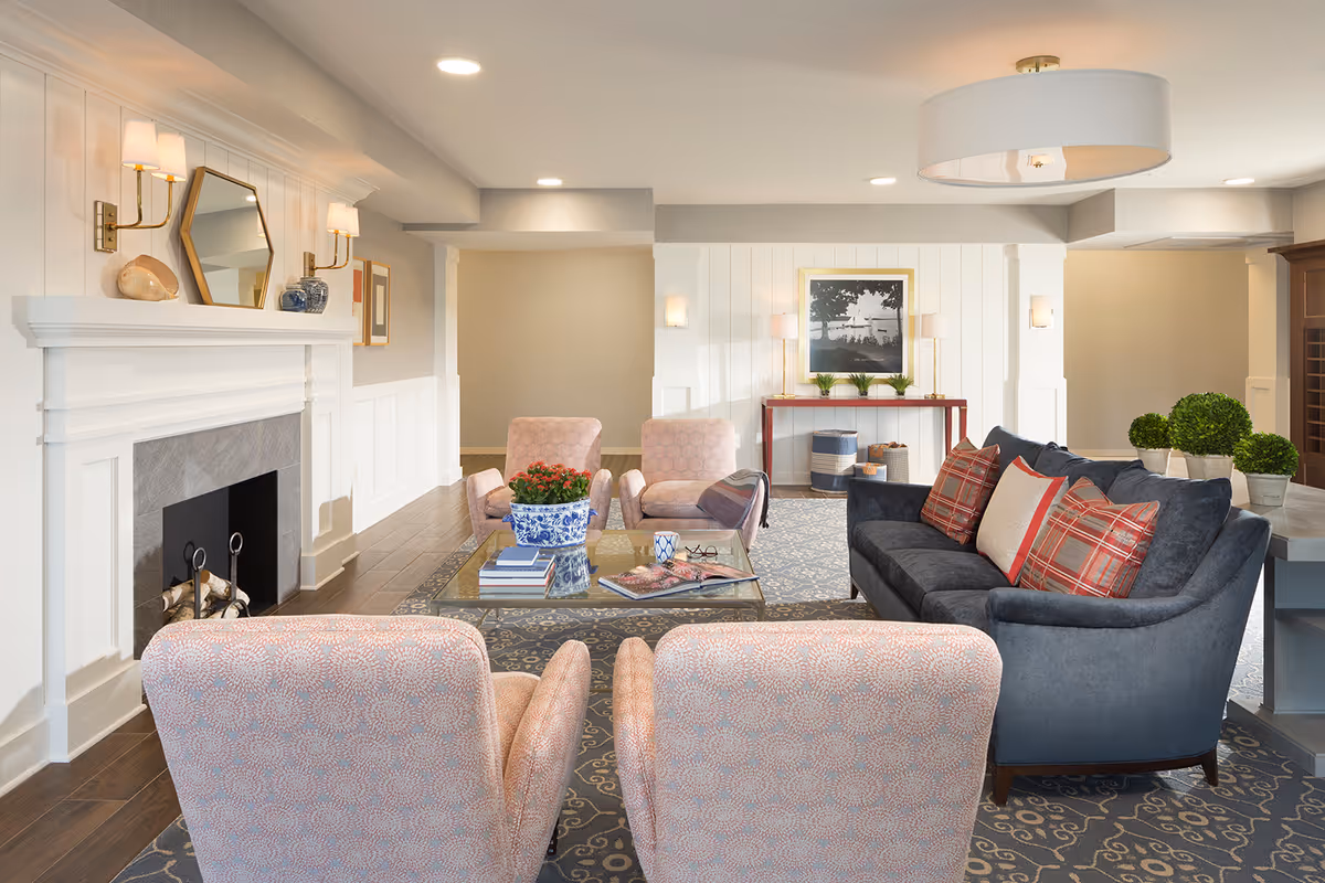 A cozy senior living facility lounge area featuring a dark blue sofa with red and beige plaid pillows, four light pink armchairs, a glass coffee table with books and a potted plant, a white fireplace with decorative items and a hexagonal mirror above it, a patterned area rug, and a console table with plants and a framed black and white photo on the wall.