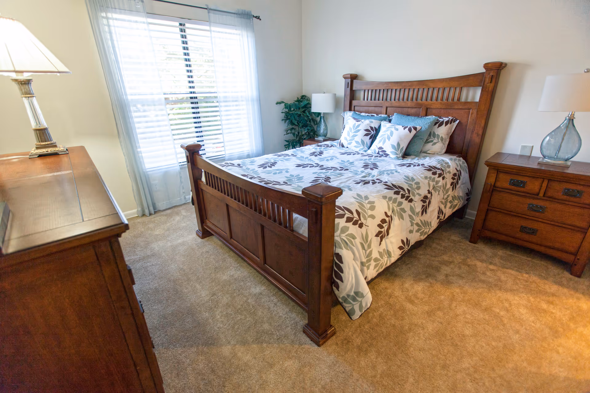 A furnished bedroom with a wooden bed frame, matching nightstands and dresser, patterned bedding, lamps, and a window with blinds and sheer curtains.