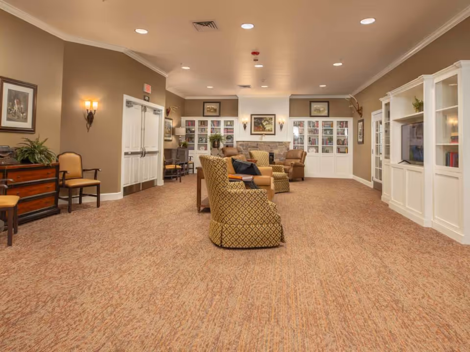 A spacious, well-lit living room area in a memory care community featuring comfortable armchairs arranged around a central space. The room has beige walls, carpeted floors, built-in white bookshelves filled with books, a TV, framed artwork, and soft wall sconces providing warm lighting.