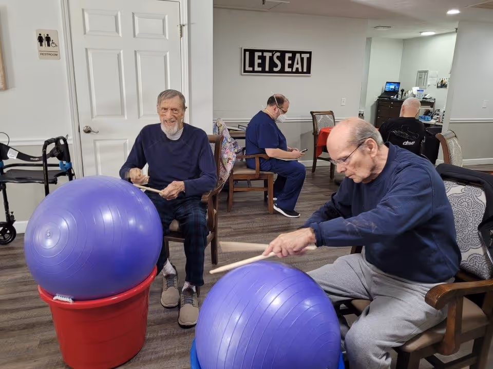 Two elderly men sitting on chairs playing drums using large purple exercise balls placed on red buckets. In the background, a man in blue scrubs is seated and looking at a phone, and another man is seated facing away. A sign on the wall reads 'LET'S EAT'. A walker is visible near a door with a restroom sign.