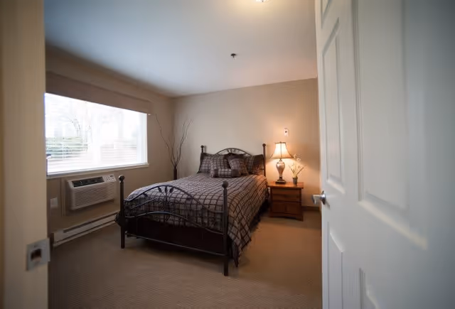 A cozy bedroom with a metal frame bed covered in a checkered brown and beige comforter and pillows. There is a wooden nightstand with a lamp and a small plant next to the bed. A large window with blinds lets in natural light, and an air conditioning unit is installed below the window. The walls are painted beige, and the carpet is light brown.