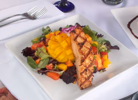 A plated dish featuring a grilled salmon fillet served on a bed of mixed greens, diced mango, tomatoes, and other fresh vegetables on a white square plate with a white tablecloth underneath.